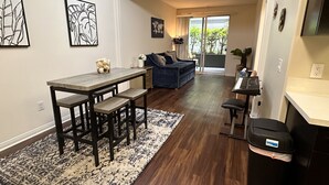 A modern living room with a dark blue sofa, a wooden dining table with benches, and a kitchen area with a black trash bin. - Lush 1 bd Koreatown w/ FREE parking & In-Unit laundry (Commerce)