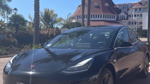 A black Tesla parked on a paved area with palm trees and a building in the background. - Lush 1 bd Koreatown w/ FREE parking & In-Unit laundry (Commerce)
