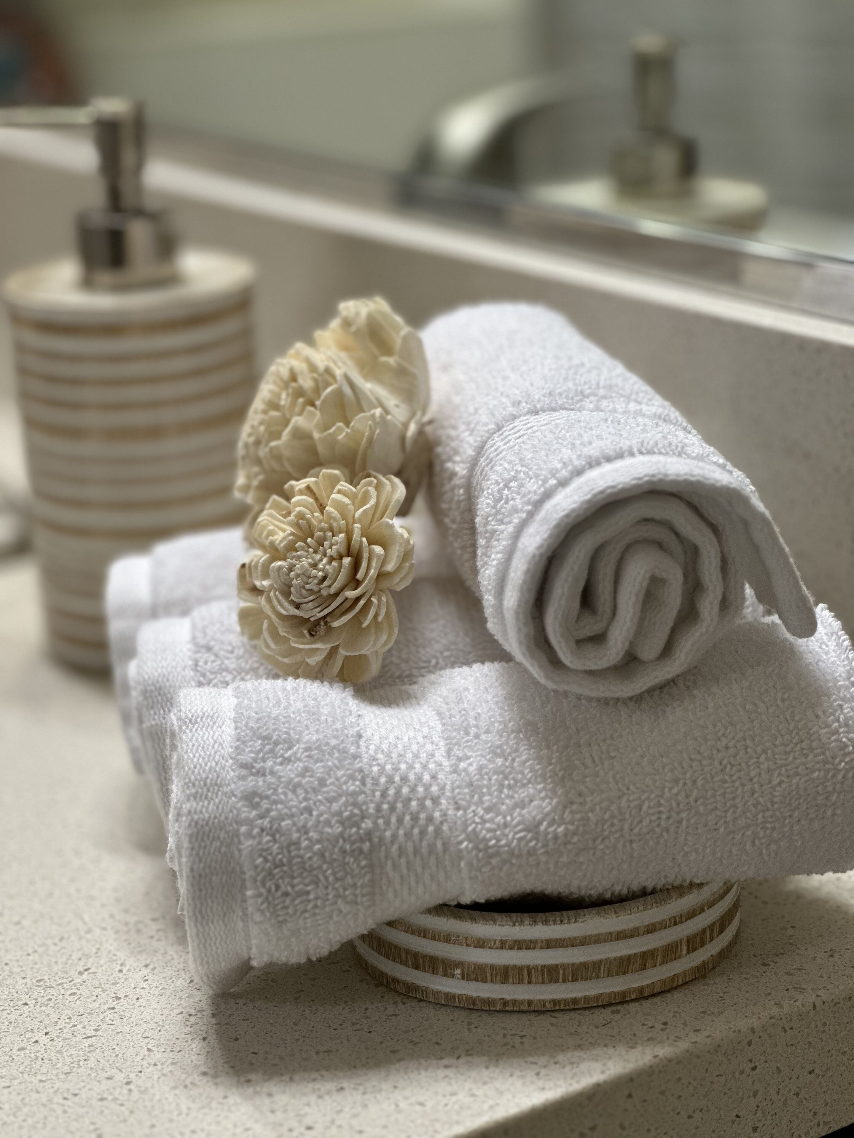 A bathroom with a towel and a decorative flower on a towel, a soap dispenser, and a mirror.