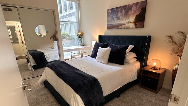 A modern bedroom with a large bed, a bedside table with a lamp, a window with a view of buildings, and a painting on the wall. - Lush 1 bd Koreatown w/ FREE parking & In-Unit laundry (Commerce)