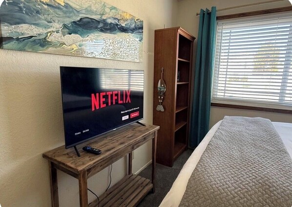 A room with a television on a wooden stand, a bookshelf, a bed with a quilt, and a window with blinds.