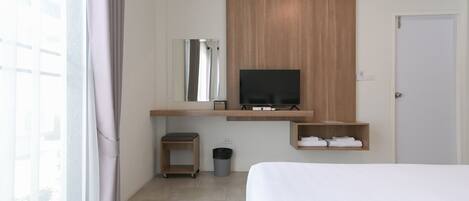 A modern hotel room with a bed, a TV mounted on a wooden panel, a small shelf with towels, a trash bin, and a window with curtains.