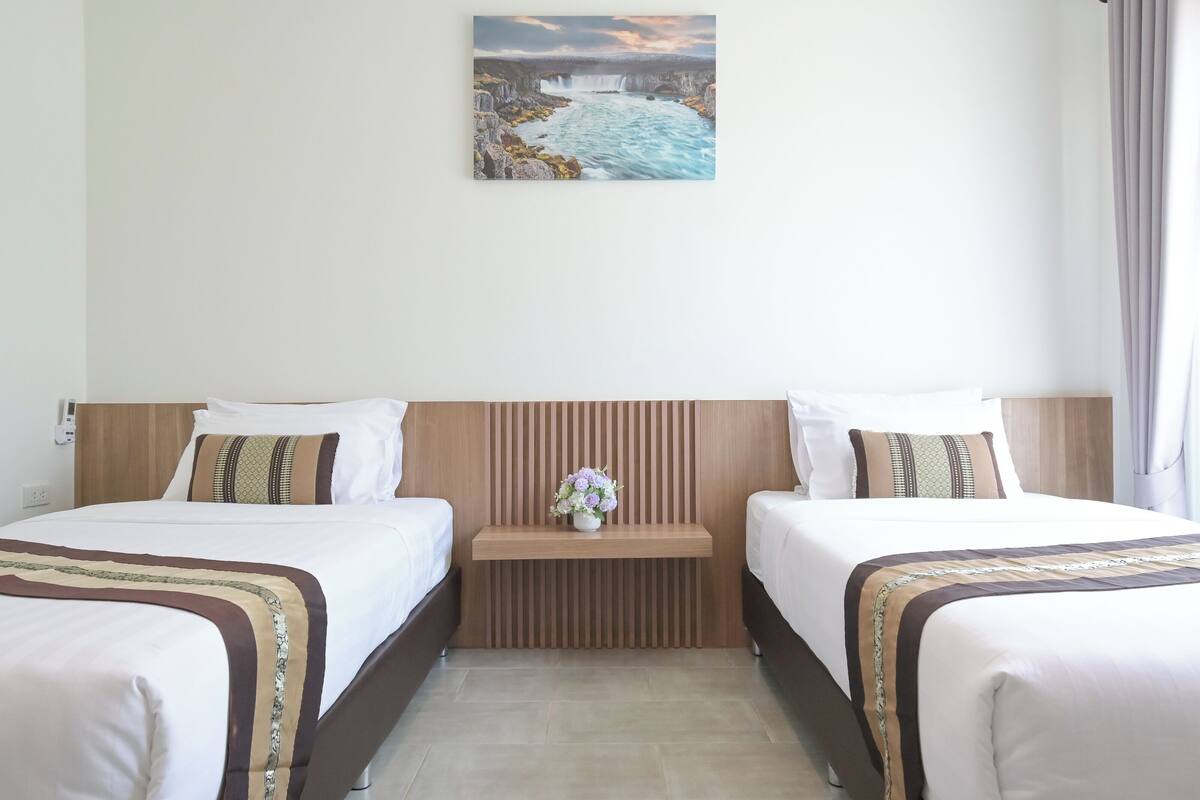 Een hotelkamer met twee bedden, een houten hoofdeinde, een kleine tafel met een bloemenvaas, en een schilderij van een waterval aan de muur.