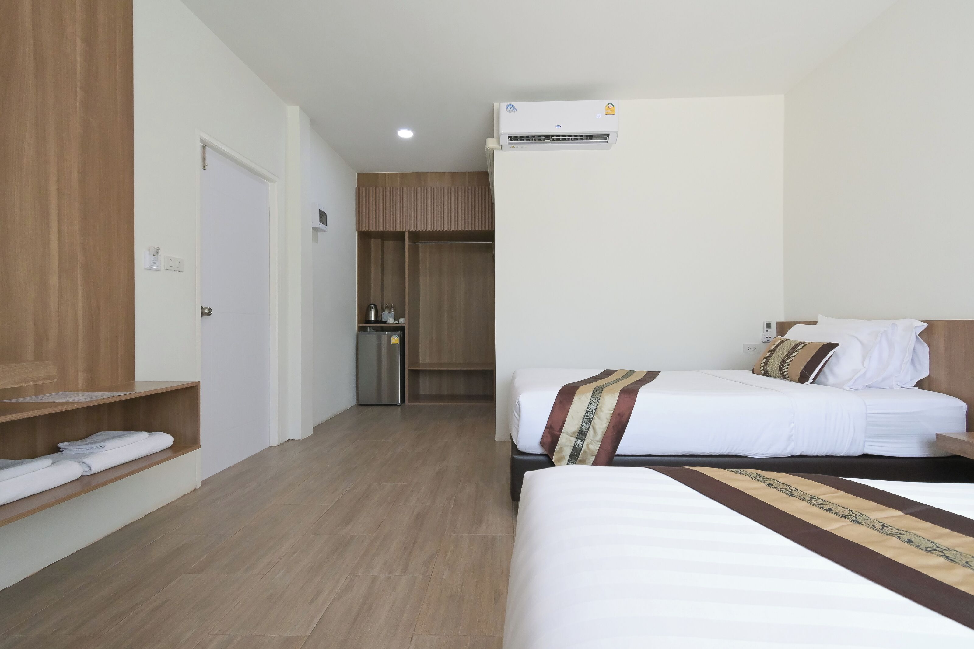 A hotel room with two beds, a wardrobe, a small kitchenette, and an air conditioning unit.