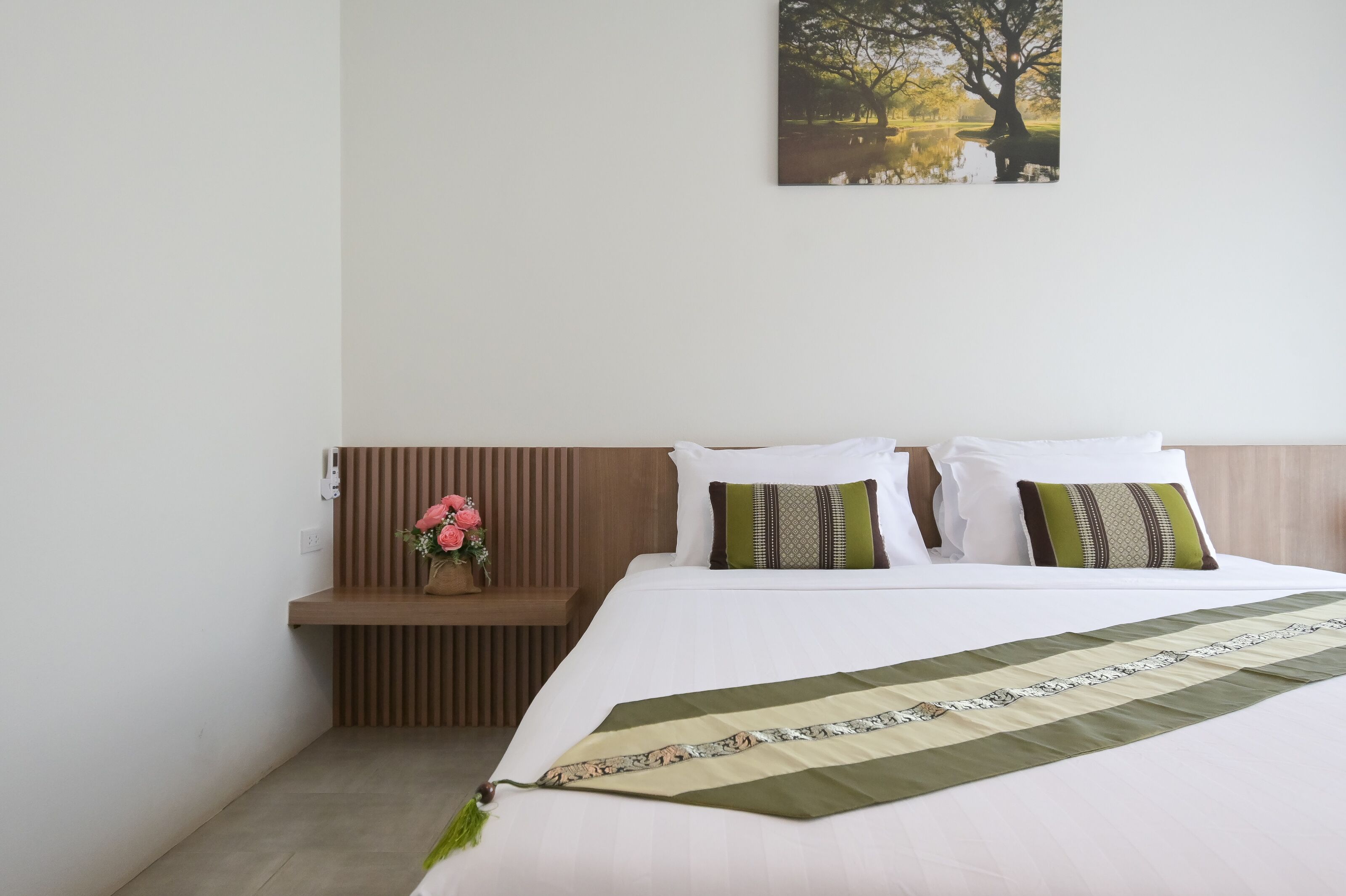 A neatly made bed with a striped blanket, a wooden bedside table with a vase of flowers, and a wall-mounted picture of a tree.