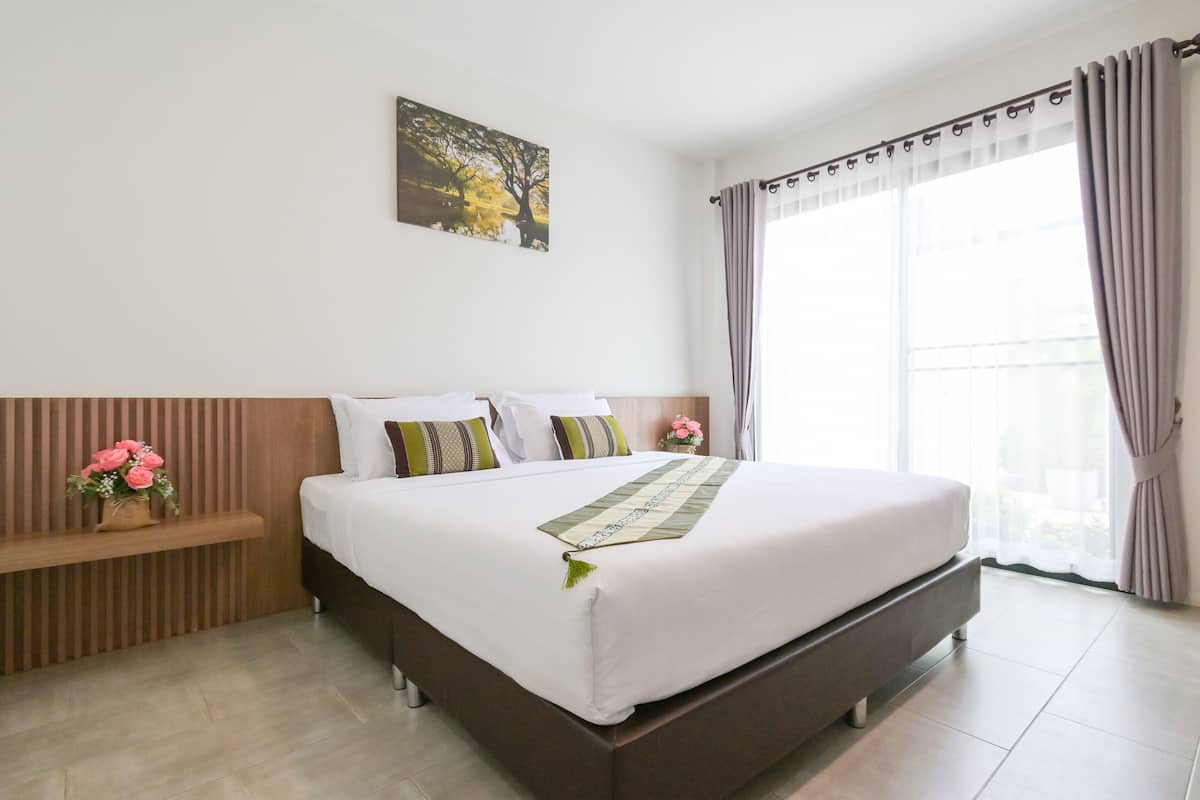 Een net opgemaakt bed met een witte deken en een decoratieve plaid, een houten hoofdbord, een klein nachtkastje met een bloevas, en een groot raam met gordijnen.