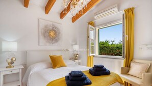 A bedroom with a bed, a bedside table with a lamp, a chair, a window with curtains, and a view of greenery outside.