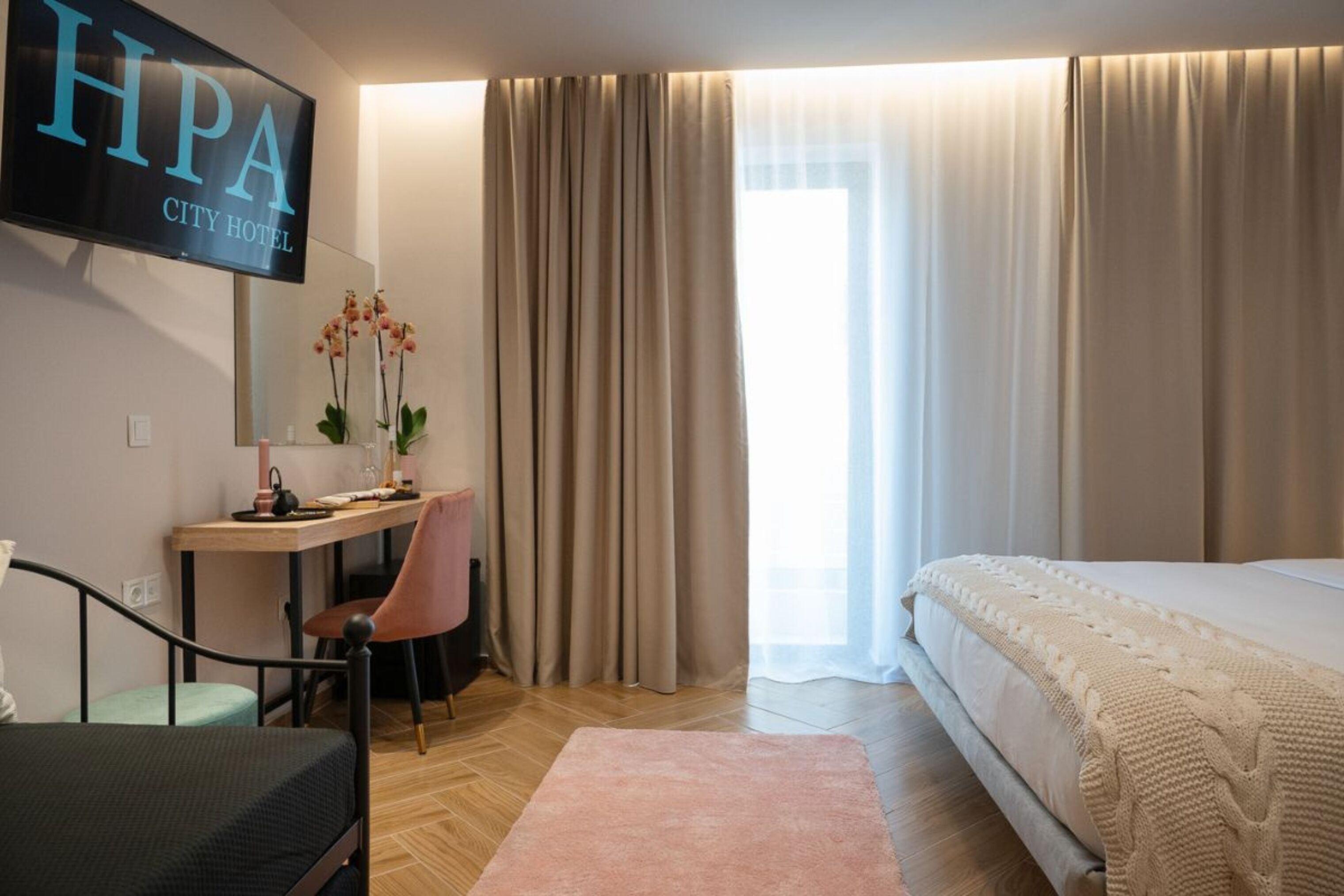 Une chambre d’hôtel comprenant un lit, un bureau avec une chaise, un téléviseur affichant le message « HPA CITY HOTEL » et une fenêtre avec des rideaux.