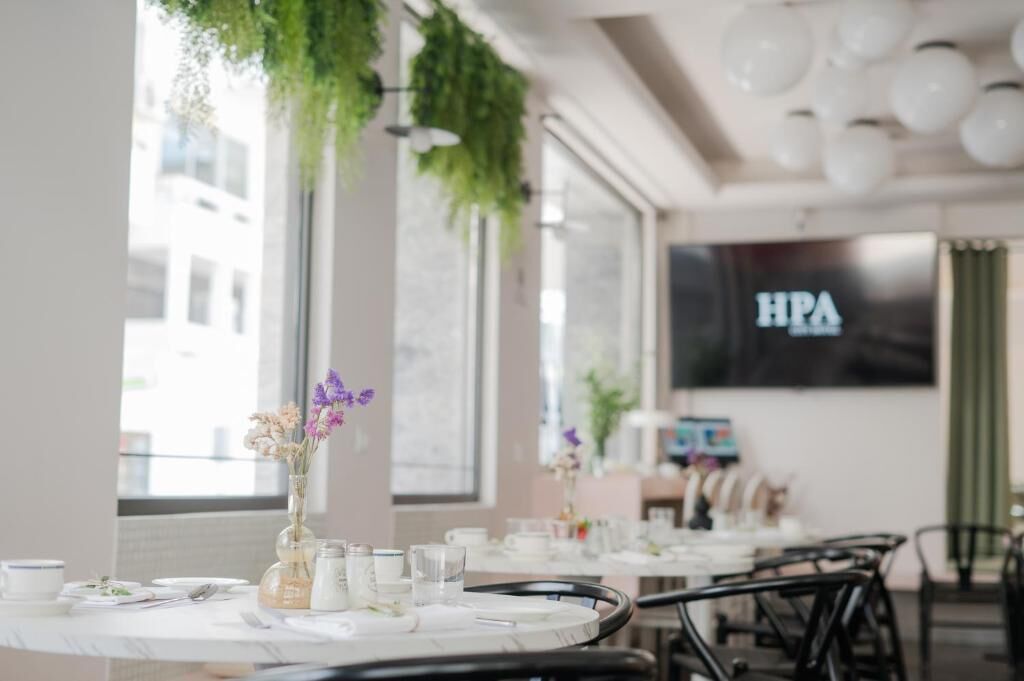A restaurant with a long table set for dining, large windows, and hanging greenery.