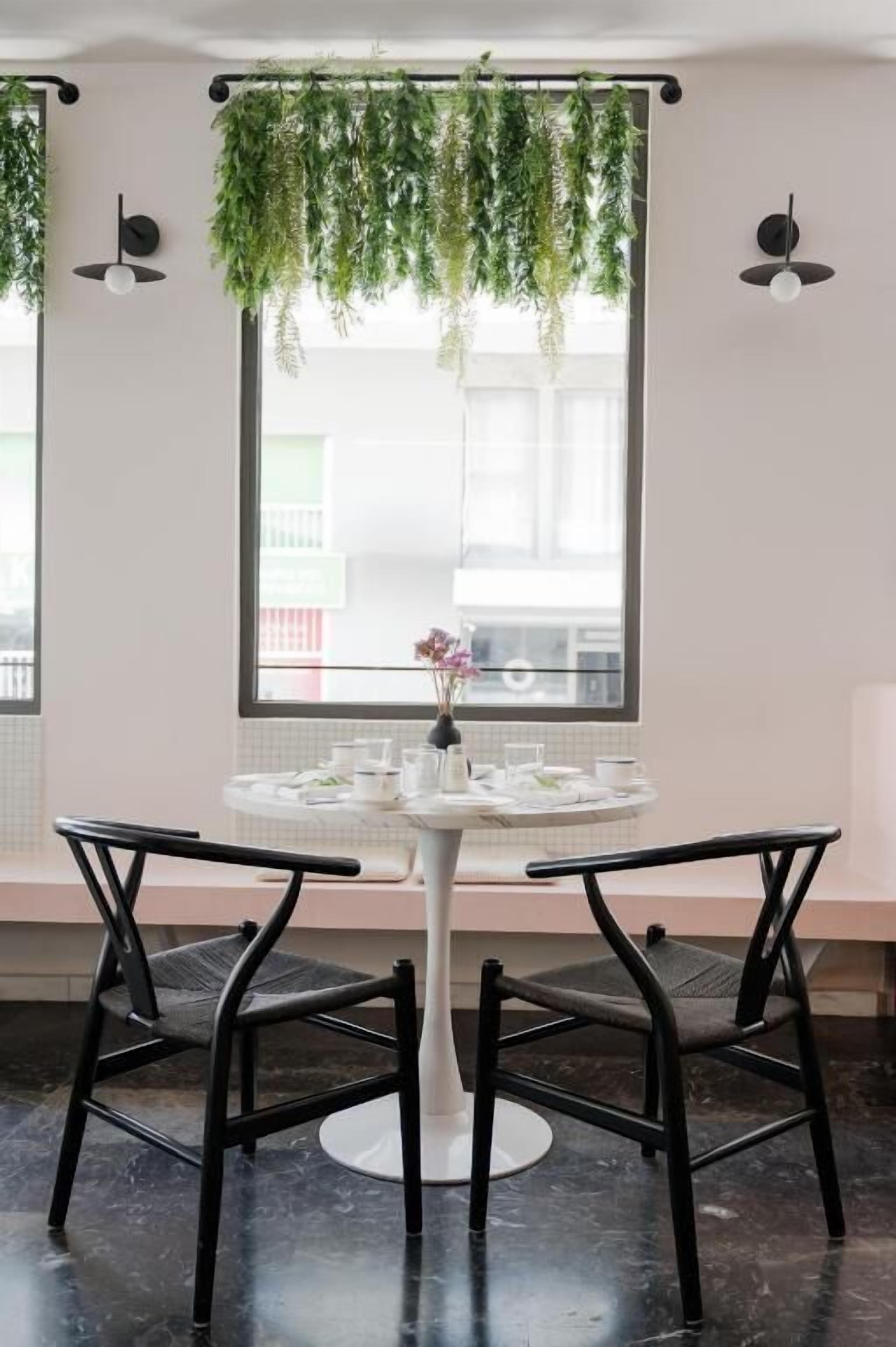 A dining area with a round table, two chairs, a vase with flowers, and hanging plants.