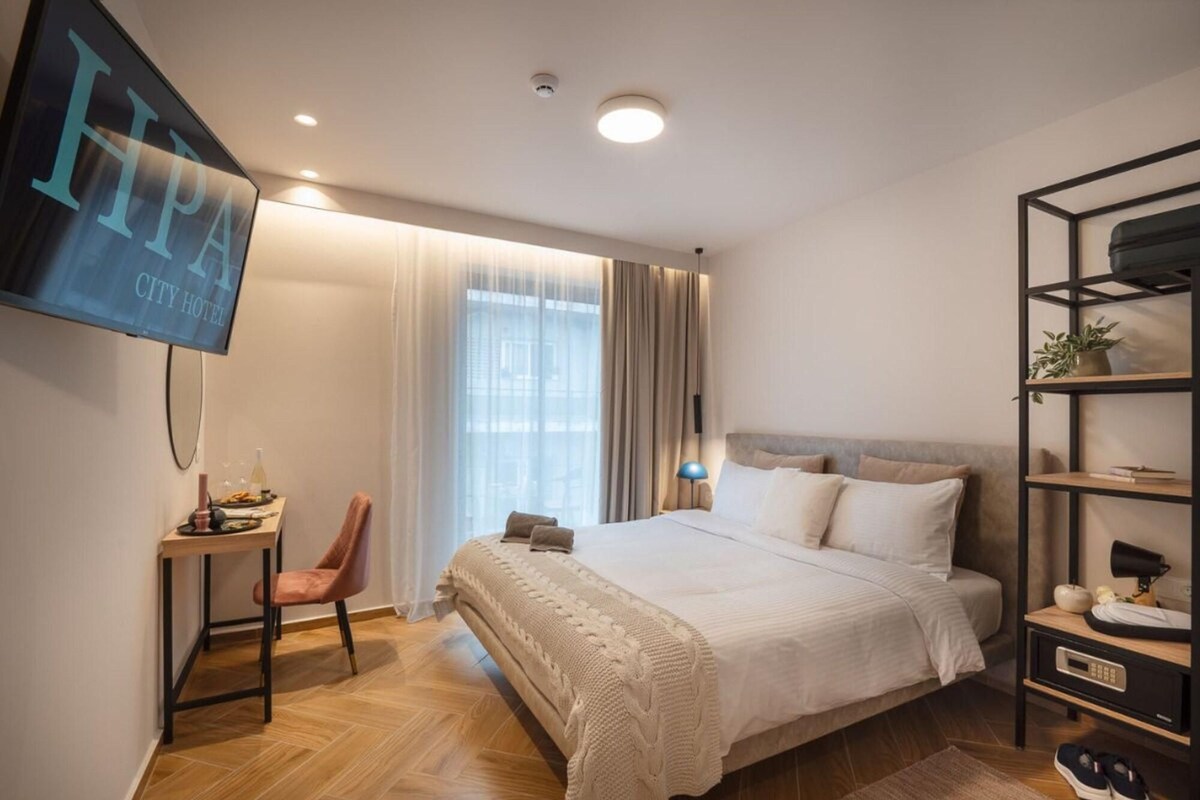 A modern hotel room with a large bed, a desk with a chair, a TV mounted on the wall, and a bookshelf.