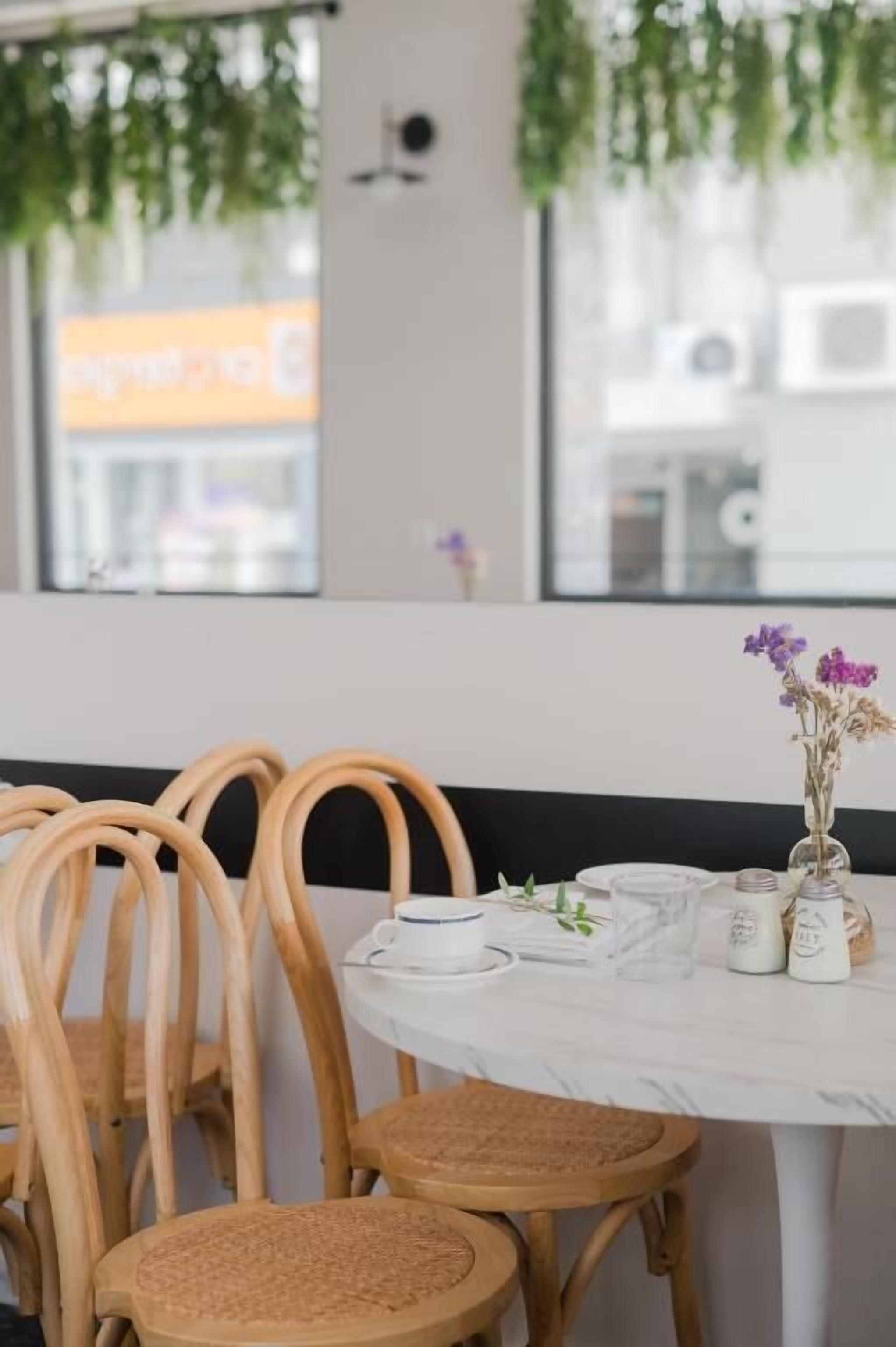 A dining area with wooden chairs and a round table, a vase with flowers, and a window with a view of the street.