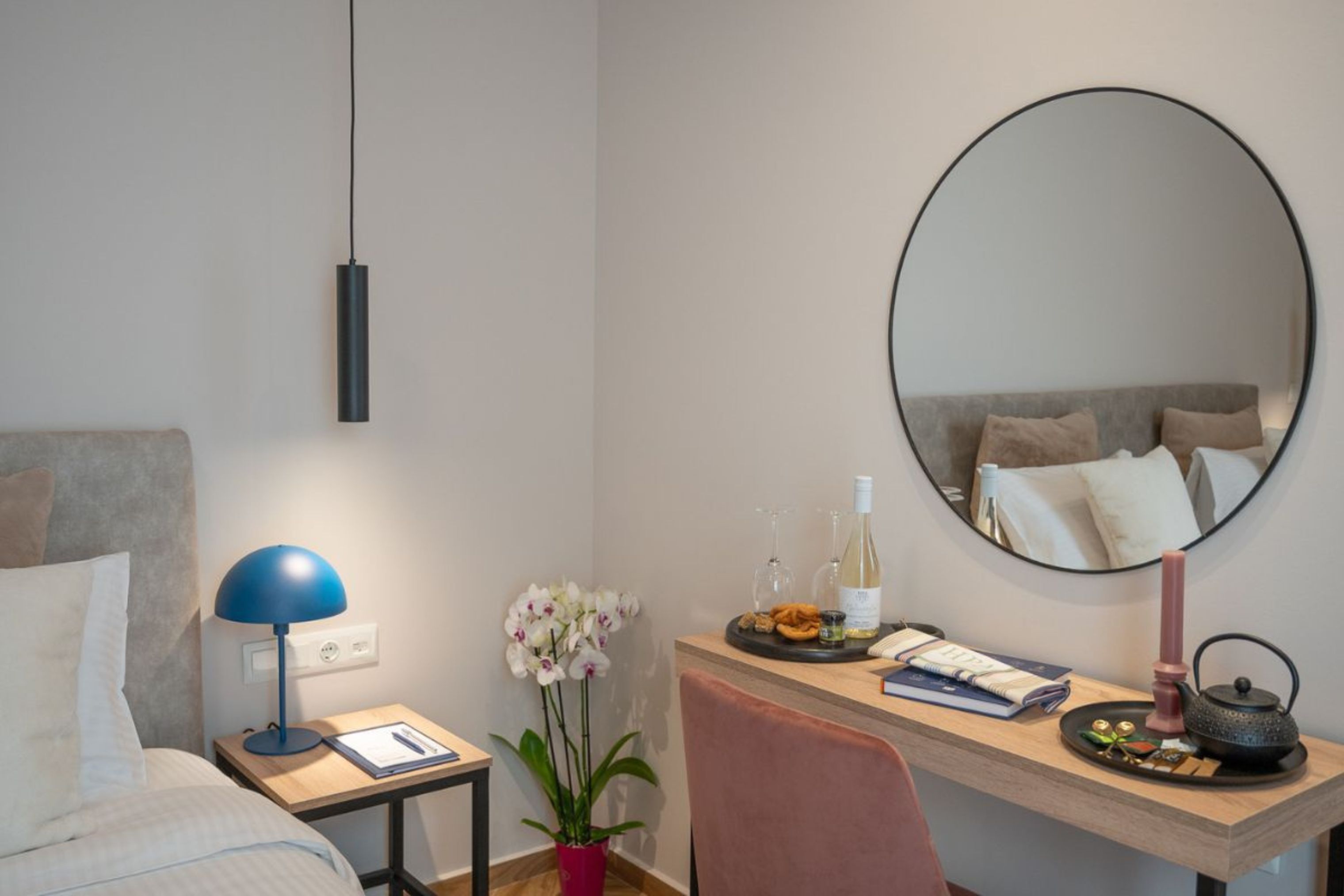 A hotel room with a bed, a desk with a lamp, a mirror, and a small table with a plant and some items.
