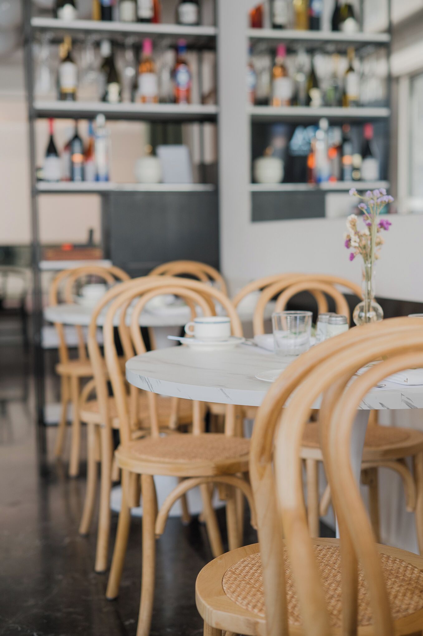 Ein Restaurantinneres mit runden Tischen und Holzsesseln, einem Tisch aus weißem Marmor und einer Theke mit verschiedenen Flaschen und Gläsern.