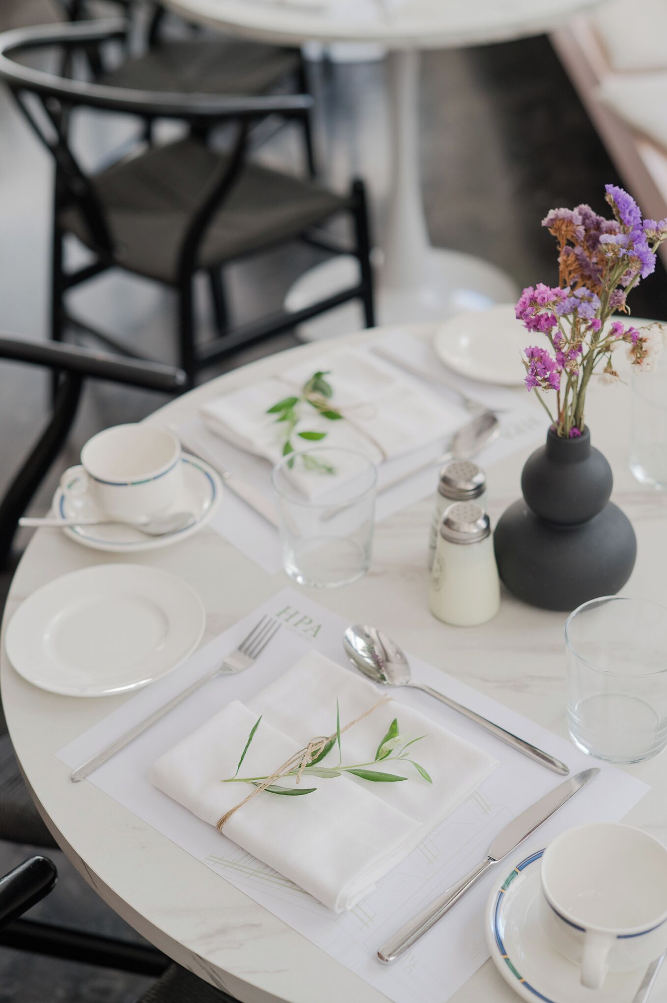Ein runder Esstisch mit weißen Tellern, Besteck und einer Vase mit lila Blumen.