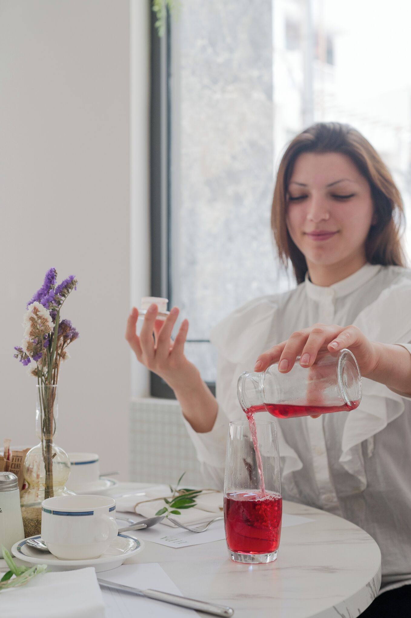 Una persona versa un liquido rosso in un bicchiere su un tavolo con un vaso di fiori e una tazza.