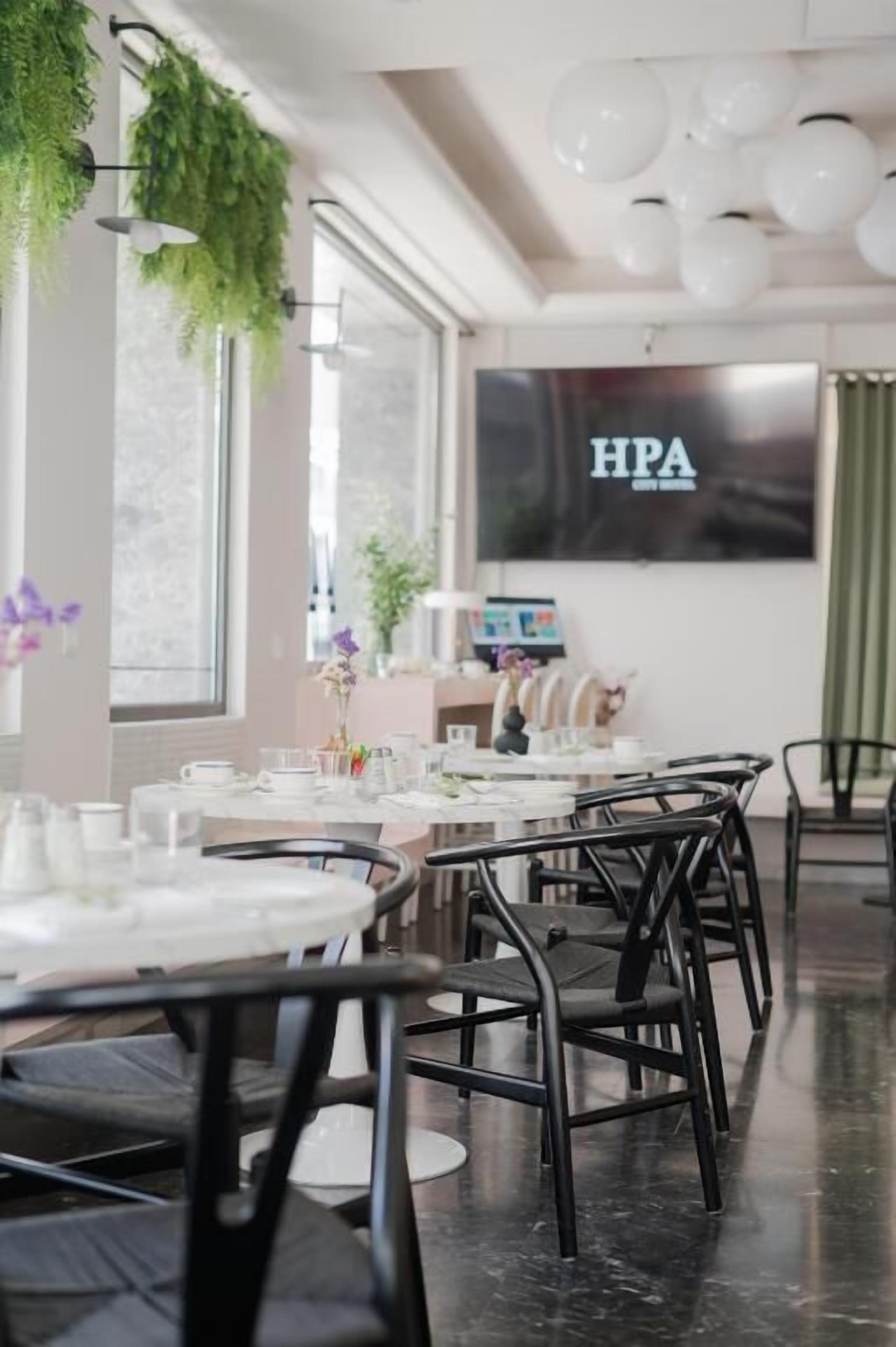 A dining area with tables and chairs, a large screen displaying 'HPA', and hanging plants.