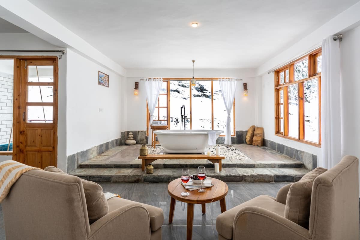 A spa room with a bathtub, two sofas, a small table with glasses and fruit, and a view of snow outside.