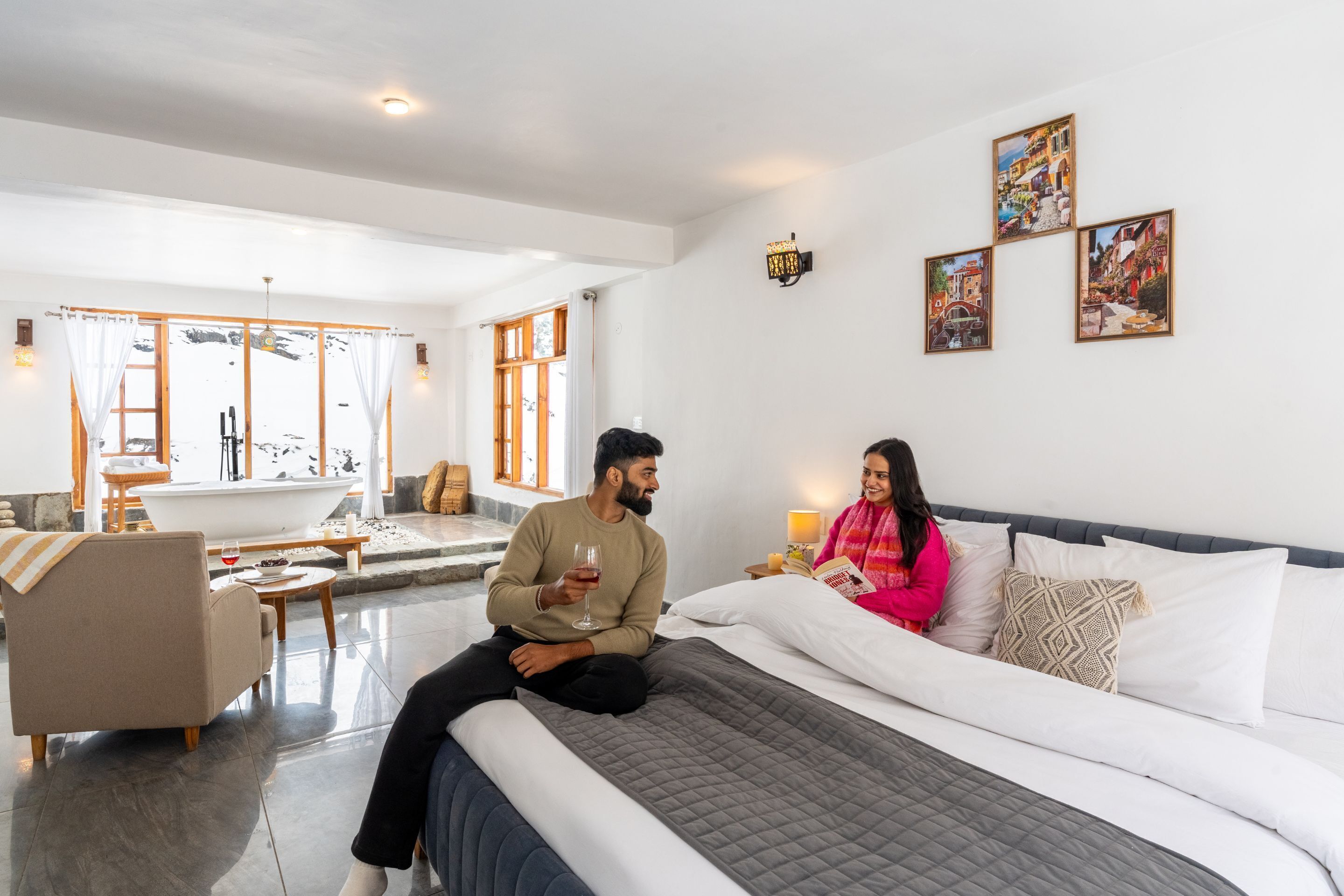 Een moderne hotelkamer met een groot bed, een zithoek met een bank en een bijzettafeltje, en uitzicht op een besneeerd landschap door grote ramen.