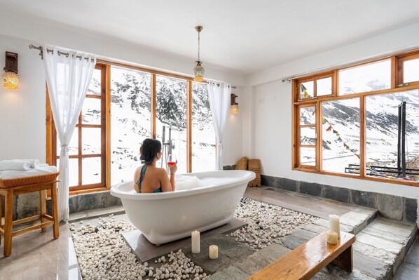 A bathroom with a freestanding bathtub, a wooden bench, and a stone floor.