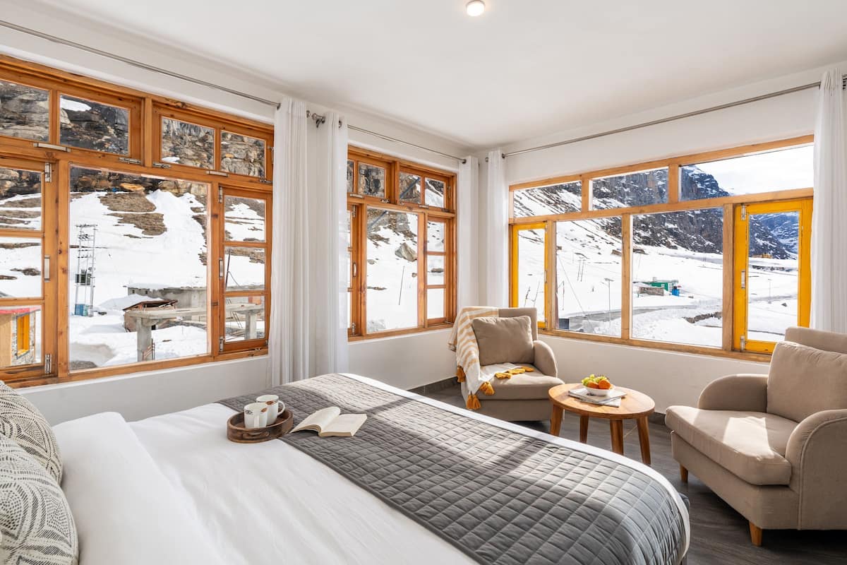 A hotel room with a large bed, two armchairs, a small table, and large windows with a snowy mountain view.