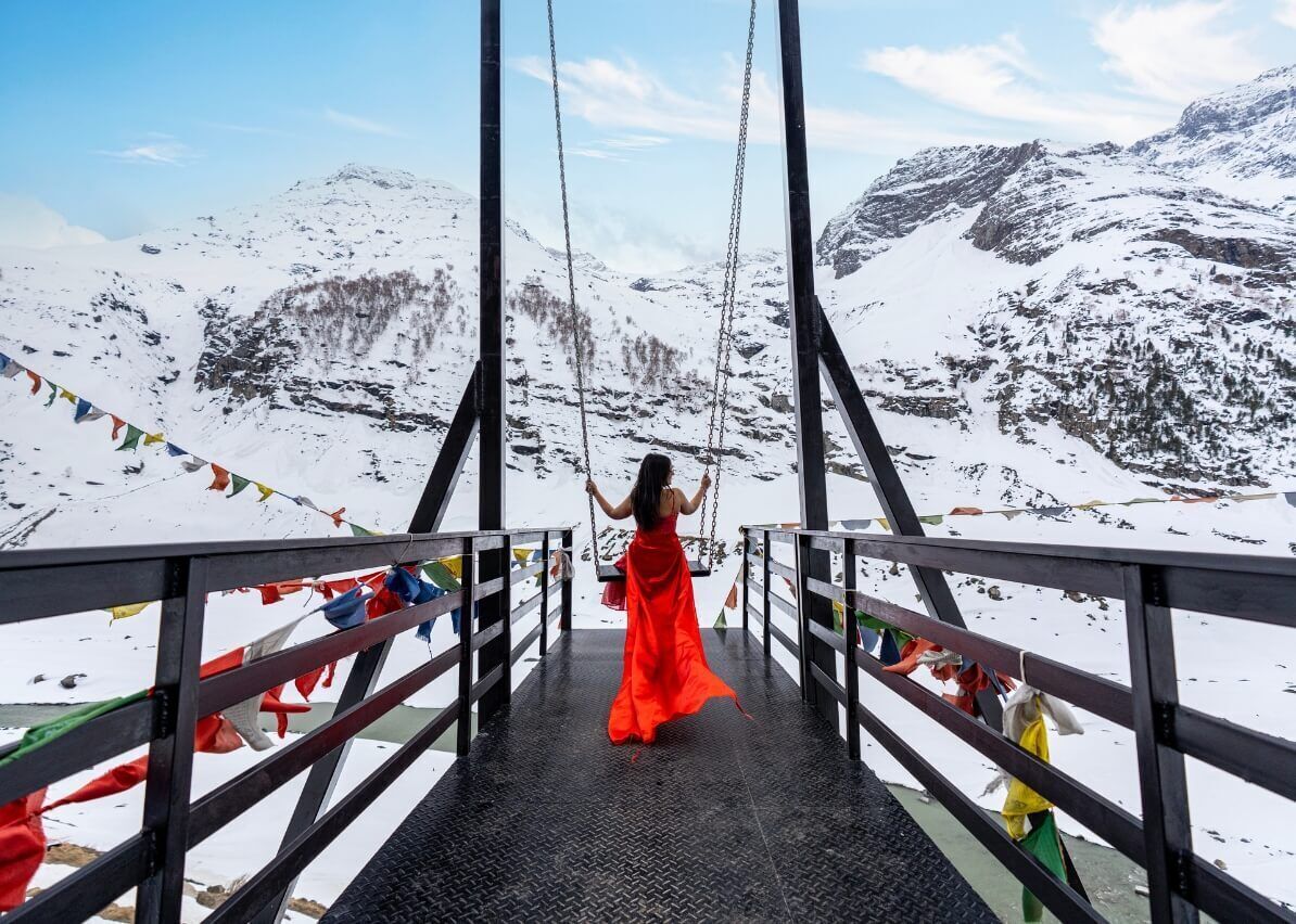 Une femme vêtue d’une robe rouge se tient sur un pont, entourée de drapeaux de prières, et contemple un paysage montagneux enneigé.
