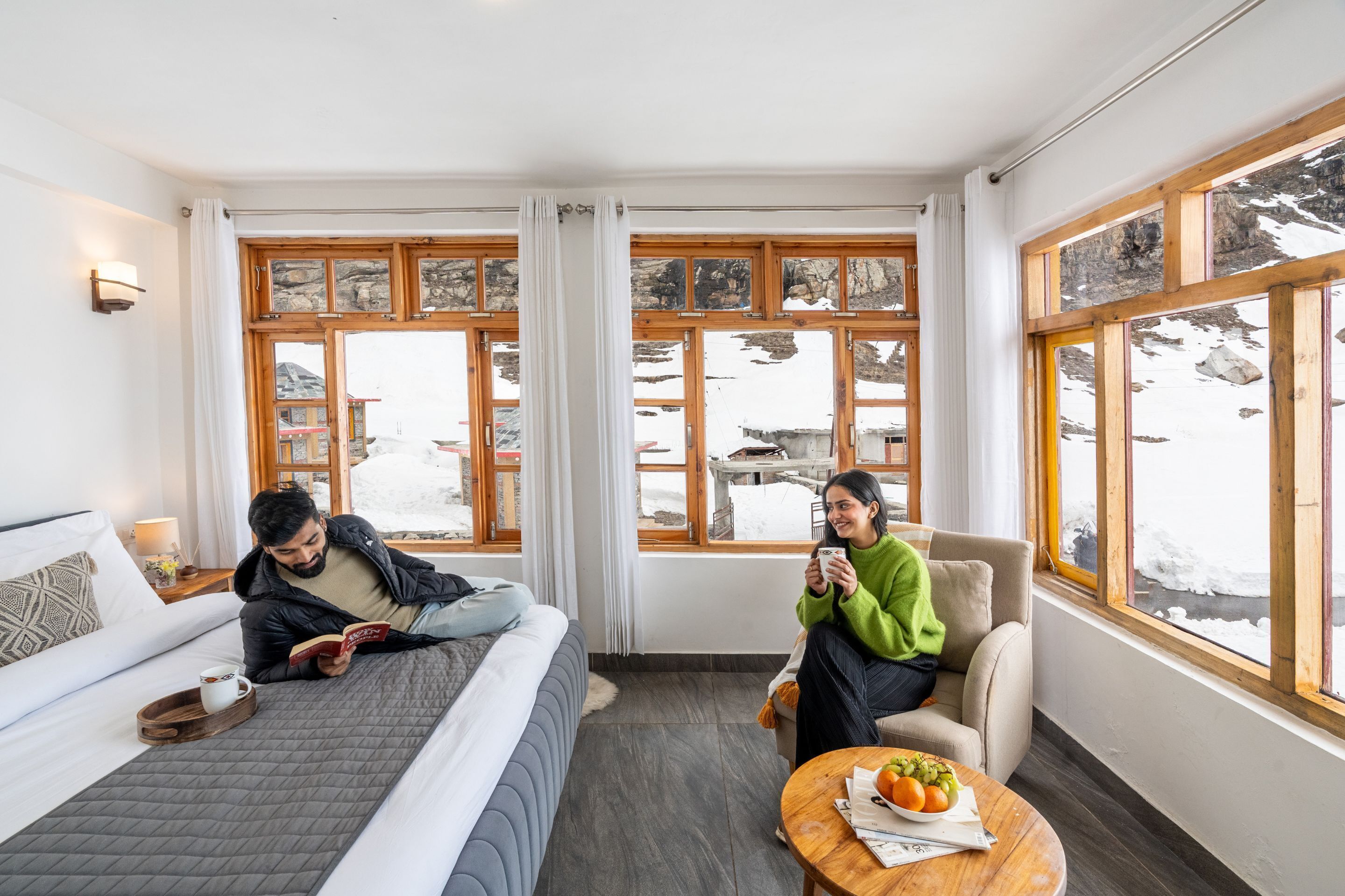 A room with a bed, a chair, a table with a fruit bowl, and large windows with a snowy view.