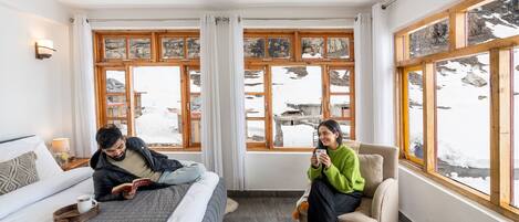 A room with a bed, a chair, a table with a fruit bowl, and large windows with a snowy view.