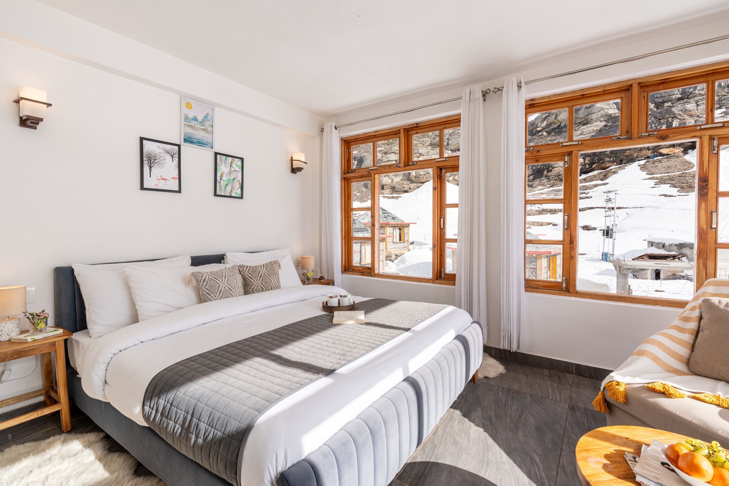 Un dormitorio con una cama grande, una mesita de noche de madera, una mesilla de noche con lámpara, una ventana con vistas a un paisaje nevado y un sofá a rayas.