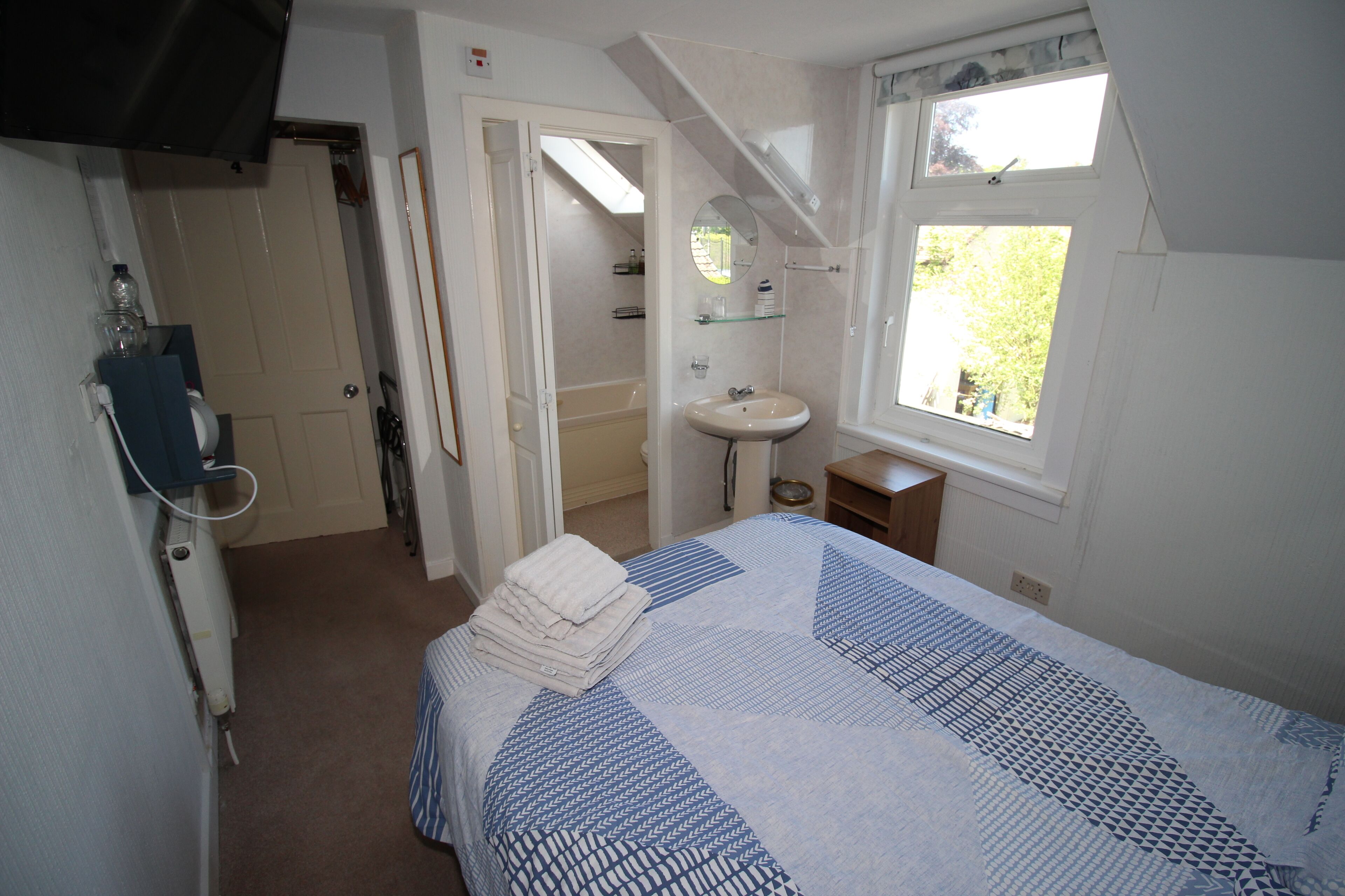 A bedroom with a bed, a window, a bathroom with a sink and mirror, and a door leading to another room.