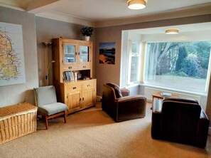 A cozy living room with a wooden cabinet, a sofa, a chair, and a large window with a view of trees. - Allandale House (Isle of Arran)