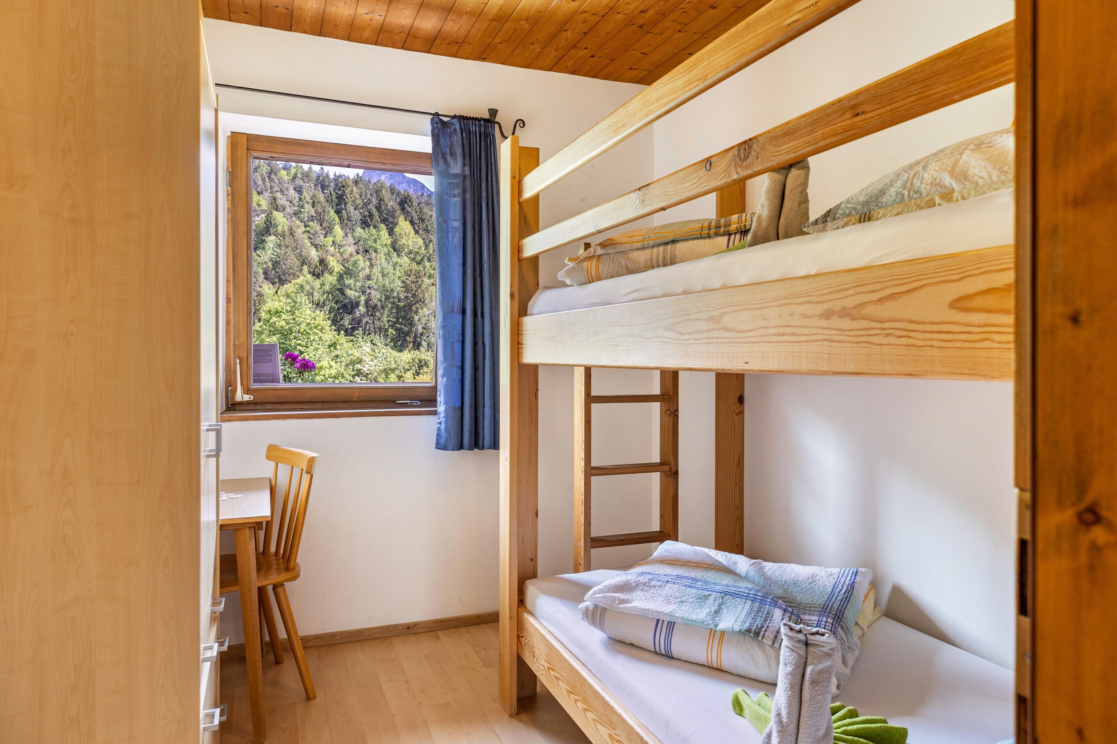 Ein Schlafraum mit Etagenbetten, einem Schreibtisch und Stuhl, einem Fenster mit Blick auf Bäume und einer Holzbalkendecke.