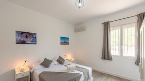 Une chambre avec deux lits, une table de chevet avec une lampe, un climatiseur fixé au mur et une fenêtre avec des rideaux.