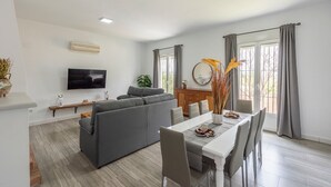 Un salon avec un canapé gris, un téléviseur, une table à manger avec des chaises et une fenêtre avec des rideaux.