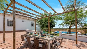 Un espace extérieur aménagé pour les repas, avec une pergola, une piscine et un bâtiment blanc doté de fenêtres.