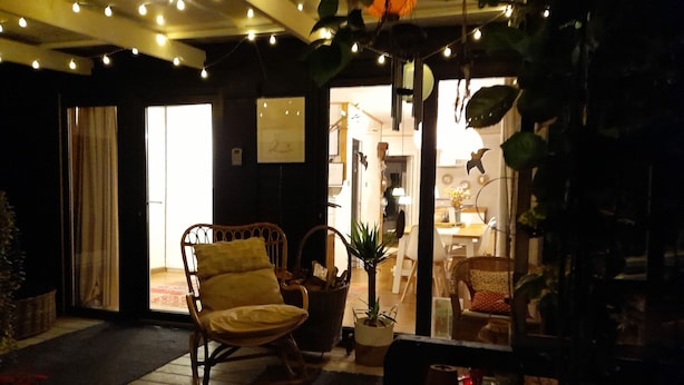 A cozy room with a wooden rocking chair, a small table, and a potted plant. There is a doorway leading to another room with a lamp and a chair.