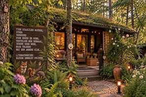 A cozy cabin with a wooden signboard, surrounded by lush greenery and a well-lit interior.