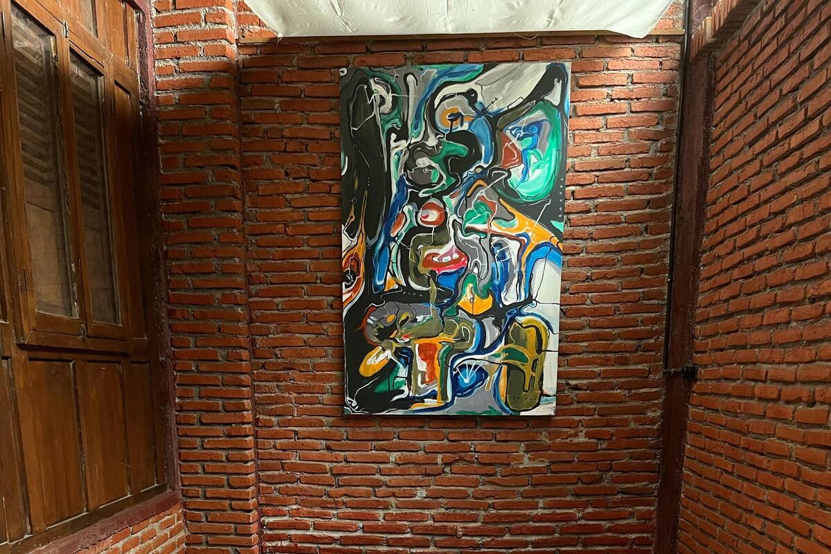 Ein schmales Zimmer mit Backsteinwand, einem Bett mit weißer Bettwäsche und einem farbenfrohen, abstrakten Gemälde an der Wand.