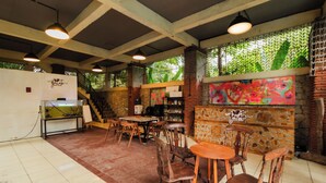 A restaurant interior with wooden tables and chairs, a stone wall with colorful artwork, and a staircase leading to an upper level. - Djawata resort & gallery  (Bangunjiwo)