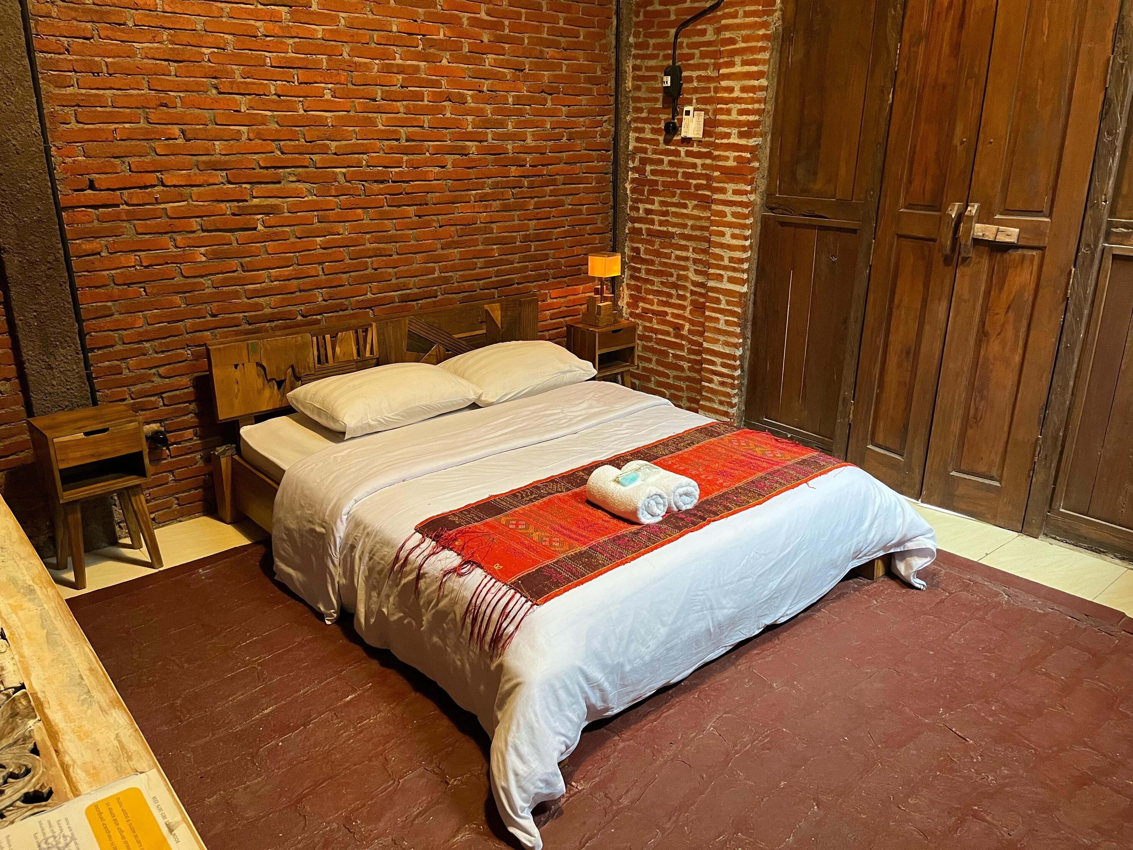 Un lit simple avec une tête de lit en bois, une couverture à motifs rouges et blancs, et une table de chevet en bois avec une lampe.