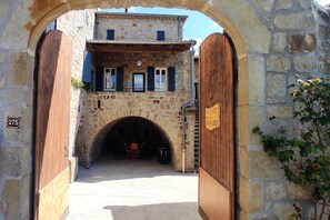 An arched stone entrance to a building with wooden doors and shutters, a balcony with a railing, and a sign on the wall.