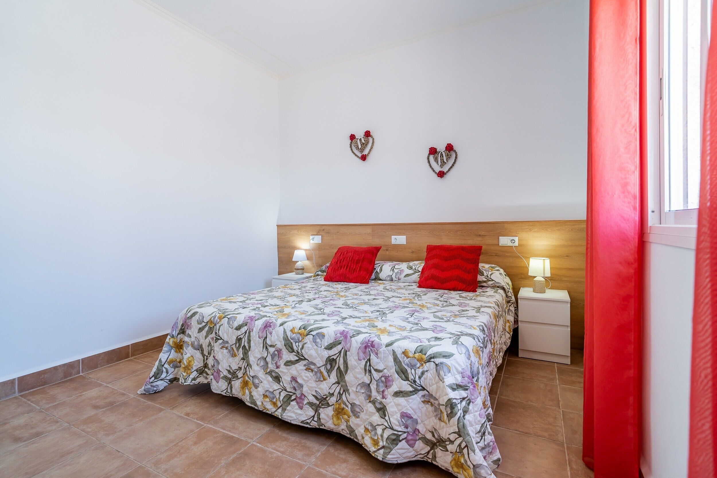 Ein Schlafzimmer mit floralem Bettbezug, zwei roten Kissen, einem hölzernen Kopfteil, zwei Nachttischen mit Lampen und Wanddekorationen in Herzform.