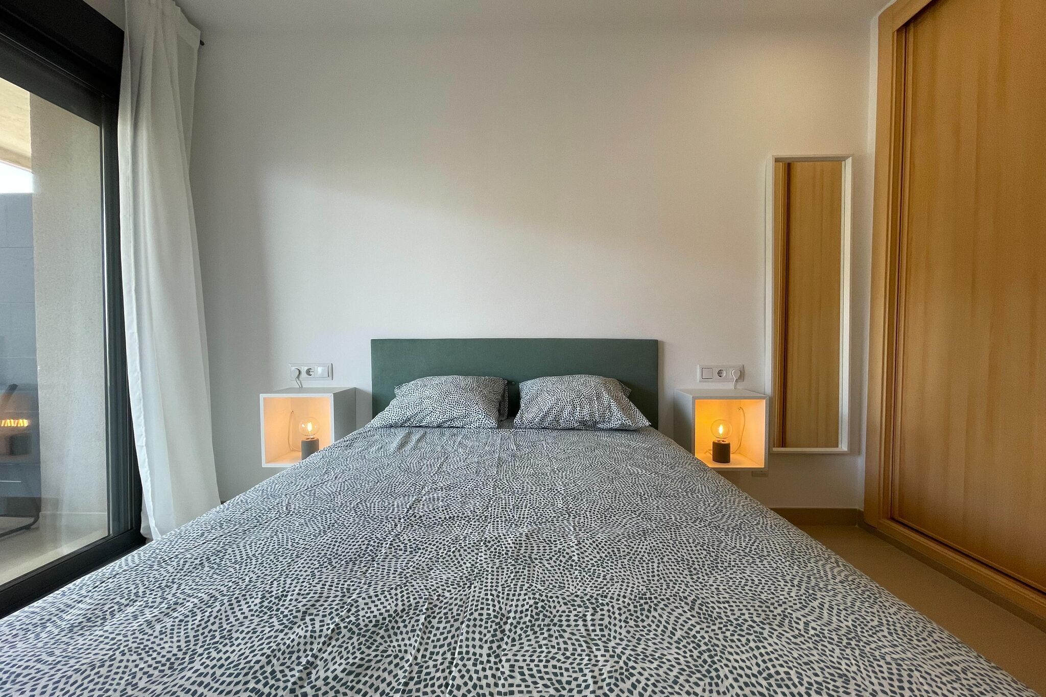 Una cama bien hecha con ropa de cama estampada, dos lámparas de noche, un ventanal con cortinas y un armario de madera.