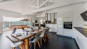 A modern kitchen with white cabinets, a wooden dining table, and a large window overlooking a deck. - Villa Sirelis with Pool and Spa (Gujan-Mestras)