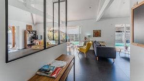 A modern living room with a large window, a sofa, a yellow chair, and a wooden table with books. - Villa Sirelis with Pool and Spa (Gujan-Mestras)