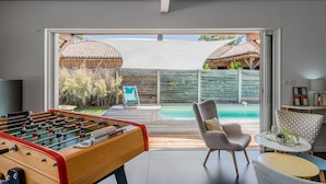 A modern indoor space with a foosball table, a pool, and a patio area with a chair and table. - Villa Sirelis with Pool and Spa (Gujan-Mestras)