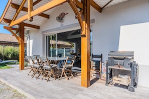 Een terras buiten met een houten pergola, een barbecue en een eettafel met stoelen.
