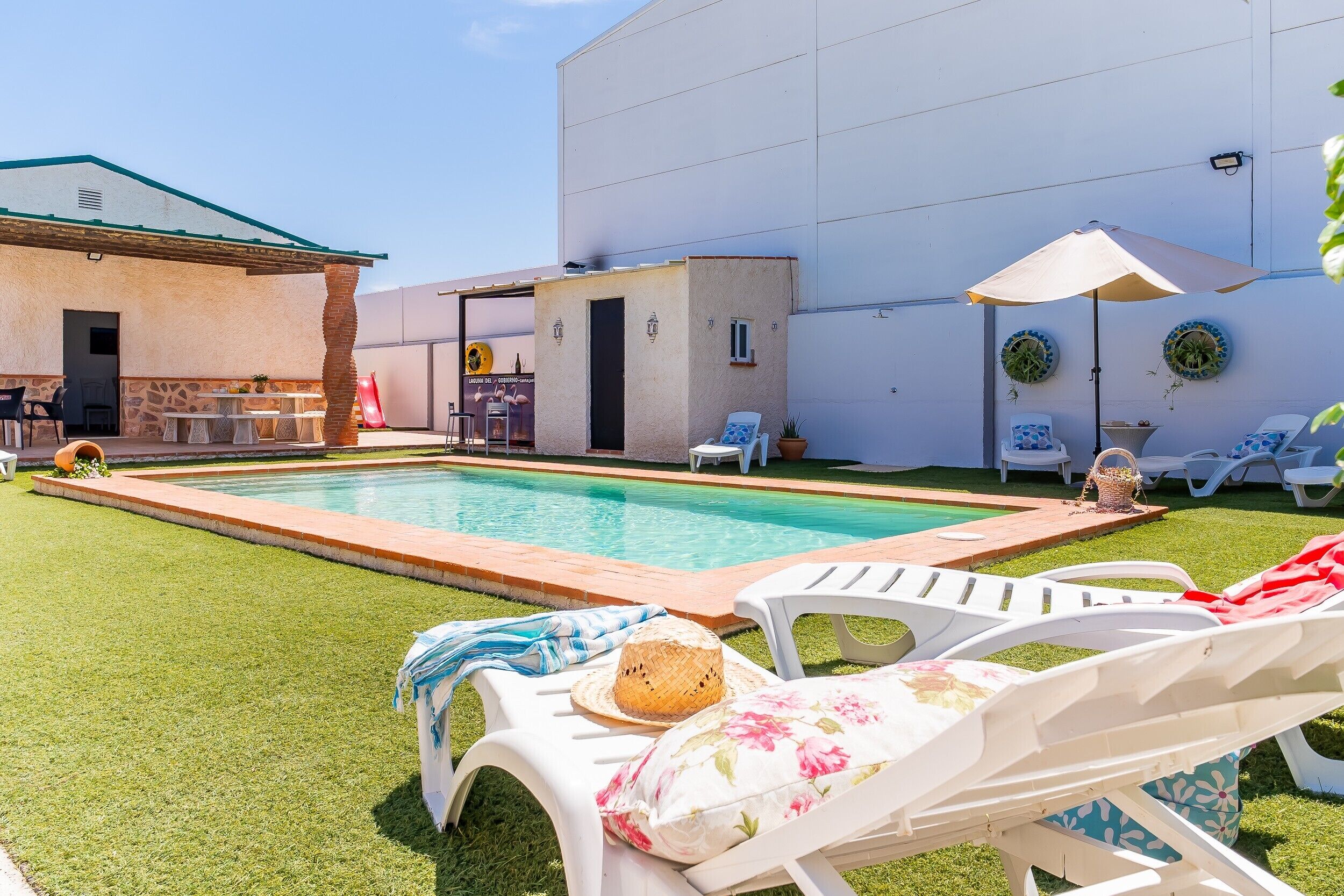 Un área de piscina con sillones de descanso, un edificio pequeño y un patio con mesa y sillas.
