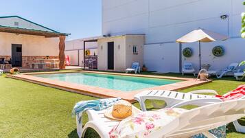 Un área de piscina con sillones de descanso, un edificio pequeño y un patio con mesa y sillas.