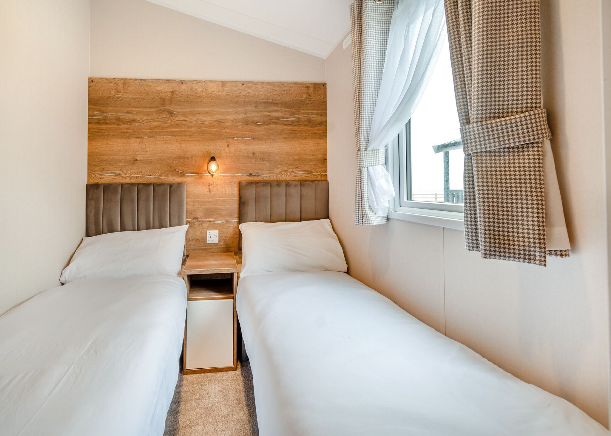 A double room with two beds, a wooden headboard, a nightstand, a wall-mounted light, and a window with curtains.